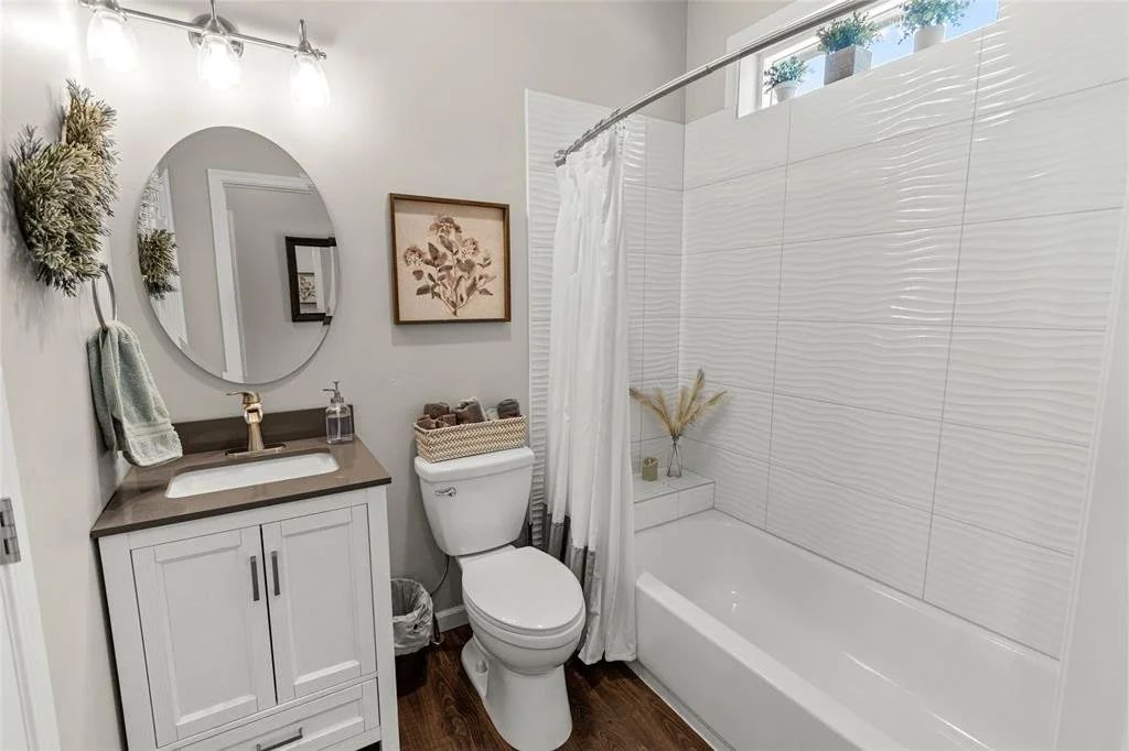 A modern bathroom with a white toilet, a vanity with a dark countertop, and a bathtub with a textured white tile wall. Decorative items include a framed botanical print, potted plants, and dried grass in a vase, with natural light coming from a small