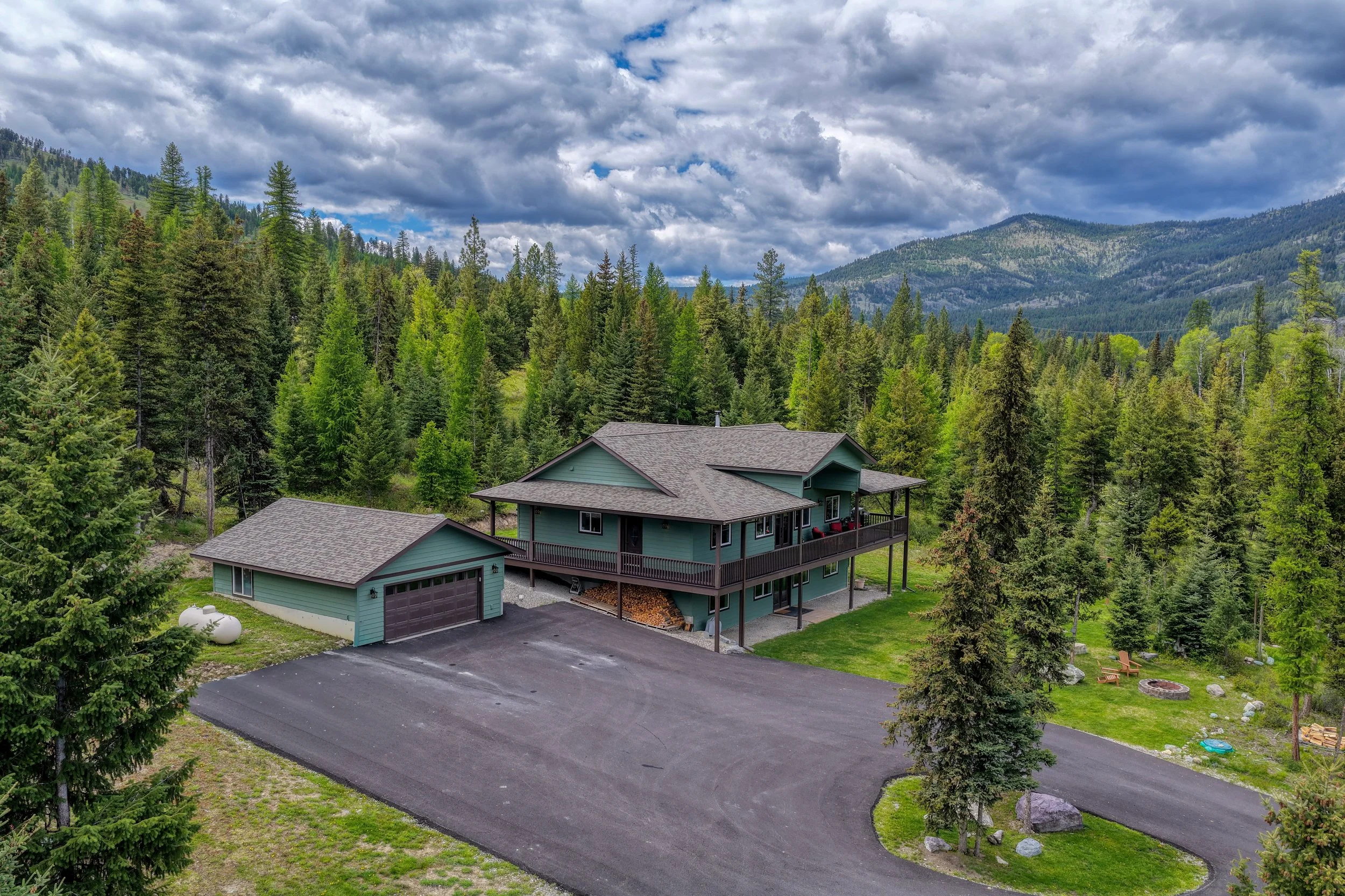 A two-story house with a dark asphalt driveway in a lush green forest with mountain hills in the background and partly cloudy sky.