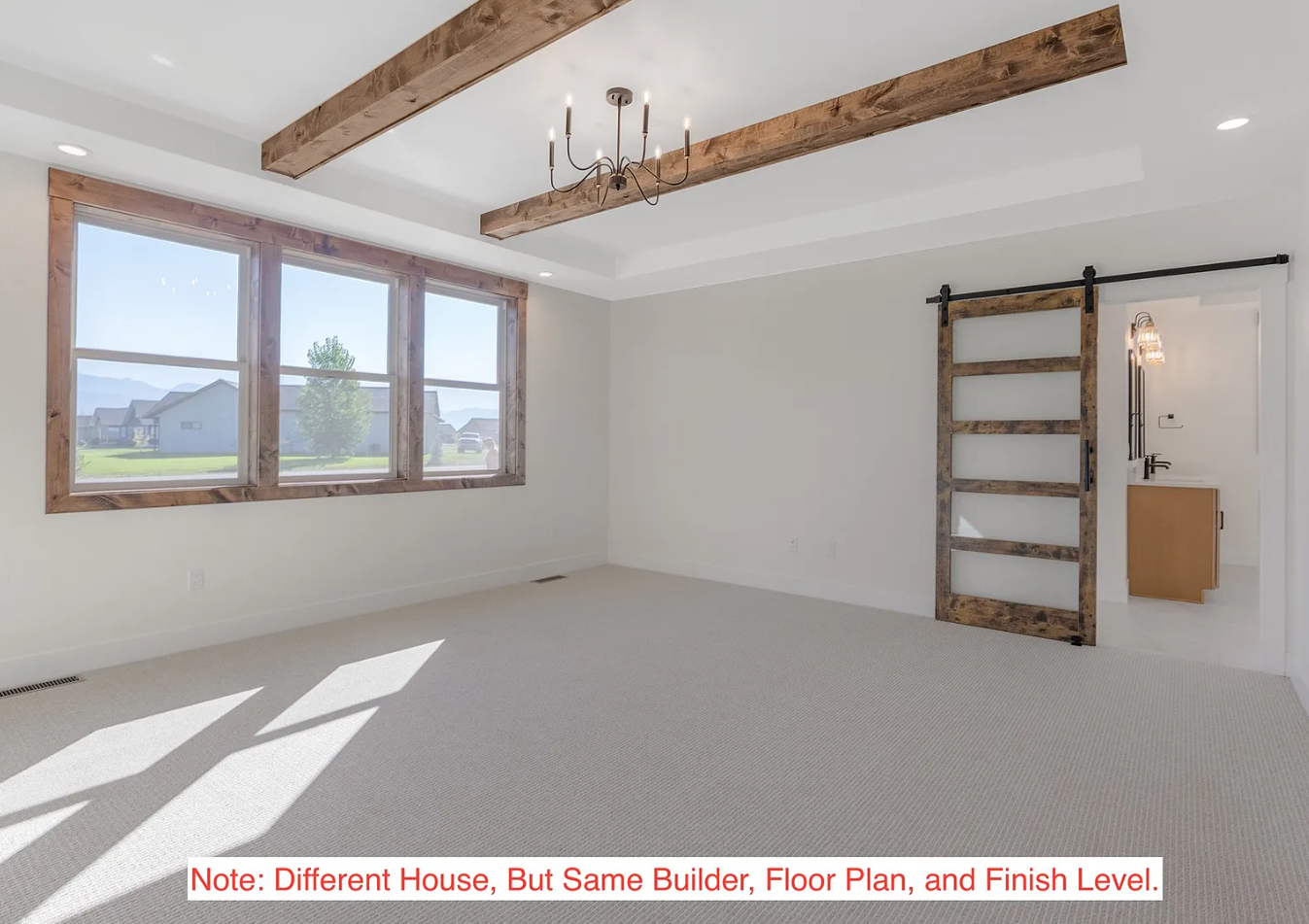 Empty room with large front window with wooden trim, beige carpet, white walls, wood beam ceiling accent, rustic wood sliding barn door on black hardware, partial bathroom with wooden vanity and wall-mounted light fixture.