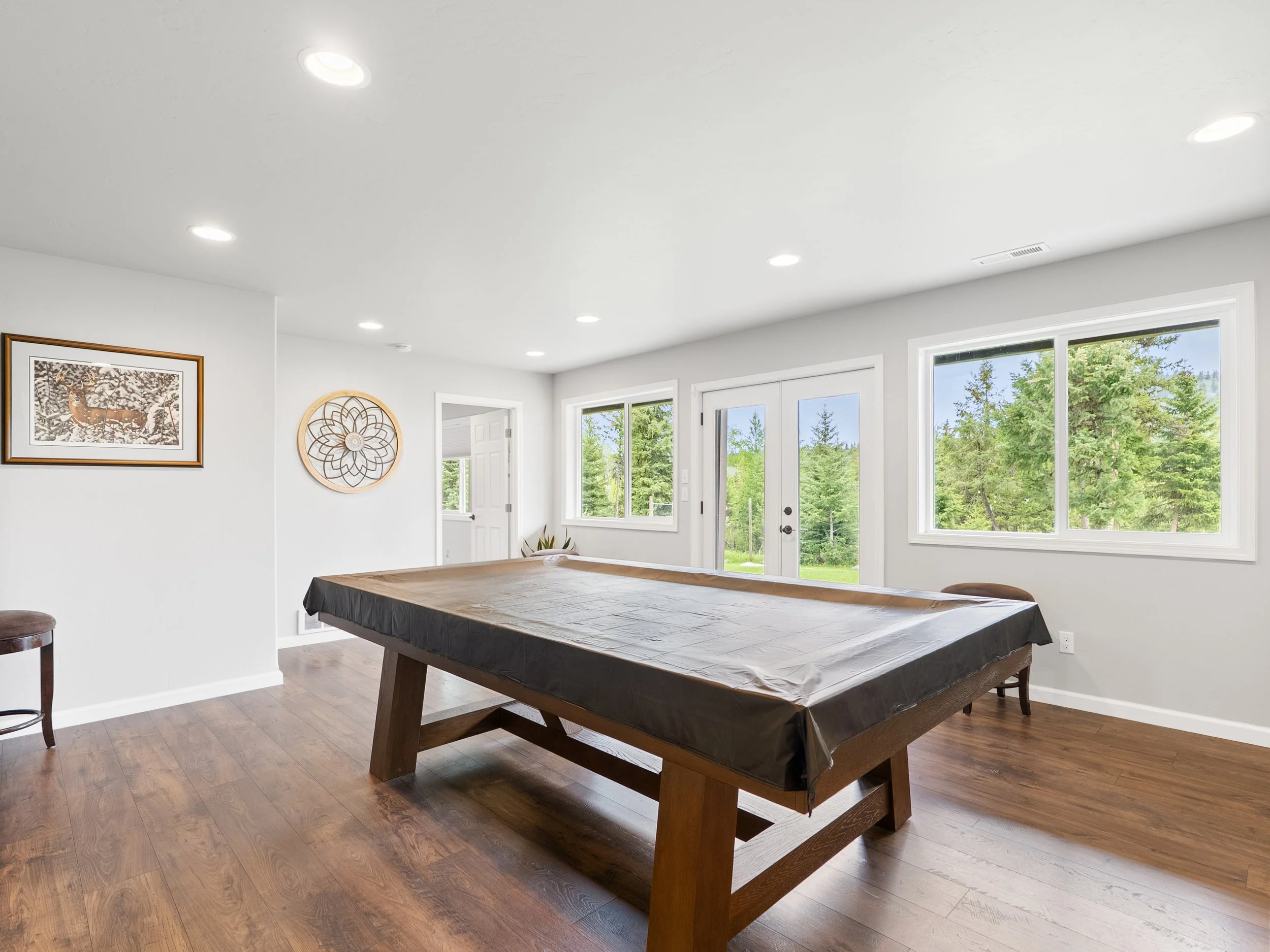 Bright living room with large windows, white walls, wooden flooring, and ceiling lights. There is a pool table covered with a black plastic cover in the center and wall decorations including a framed picture and a circular decorative piece.