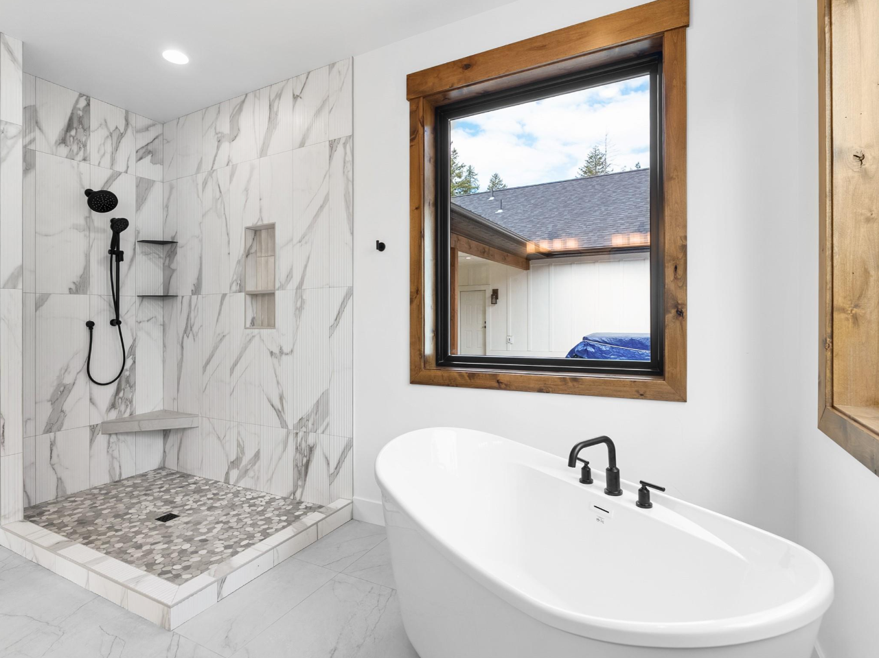 Modern bathroom with a large walk-in shower featuring white marble tiles with gray veining, a built-in niche, and pebble flooring. A freestanding bathtub with black fixtures sits next to a window with a wooden frame, showing an outdoor view of a hous