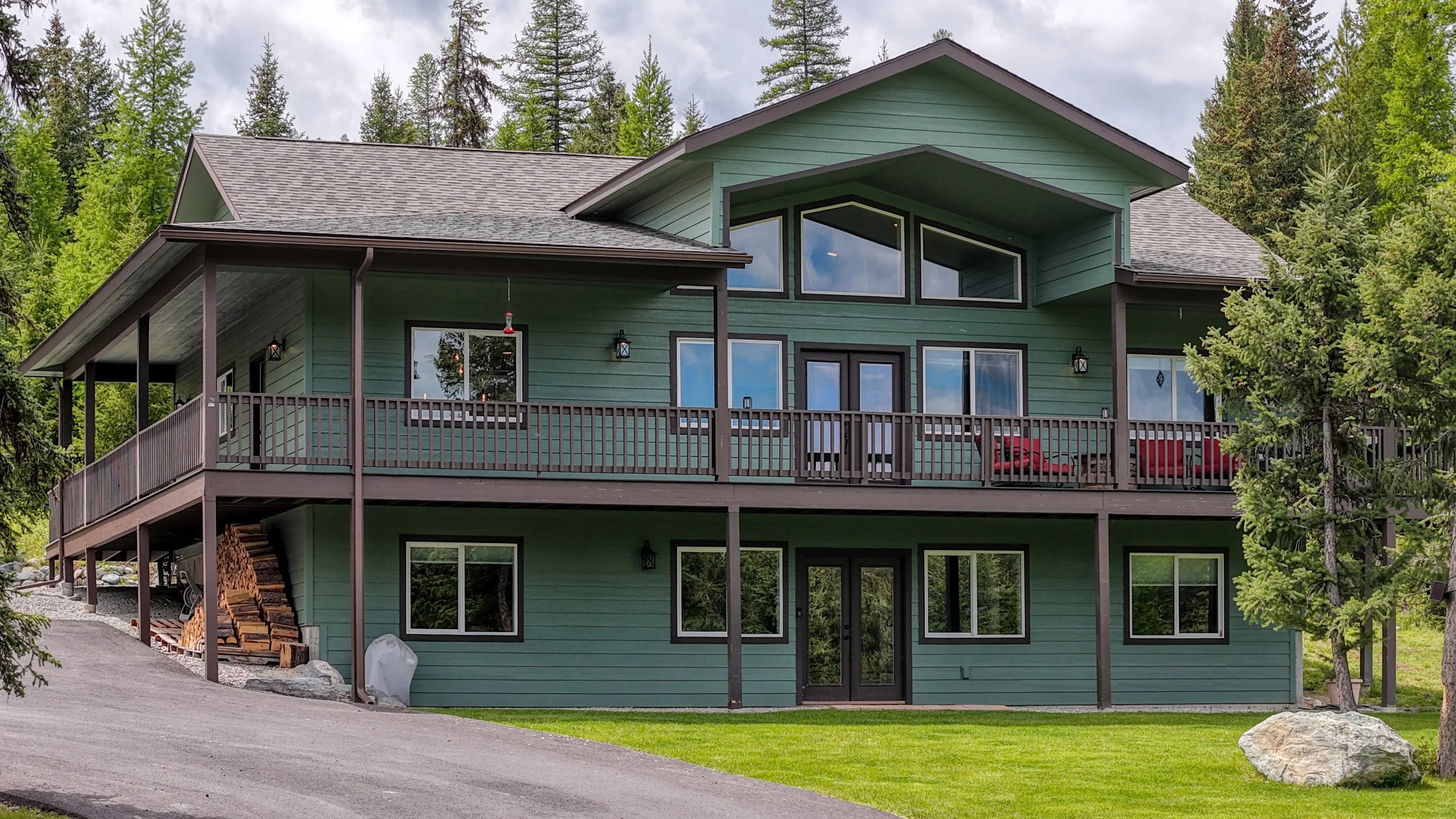 A large, multi-story house painted in green with dark trim, featuring multiple windows and a spacious balcony with outdoor furniture, set in a forested area surrounded by trees.