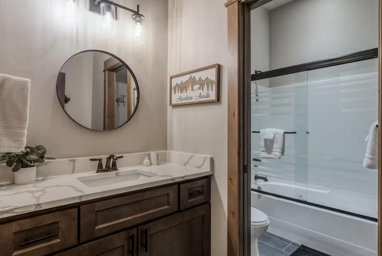 Modern bathroom with a wooden vanity, marble countertop, round mirror, and a glass-enclosed shower with towels hanging on the towel bars.