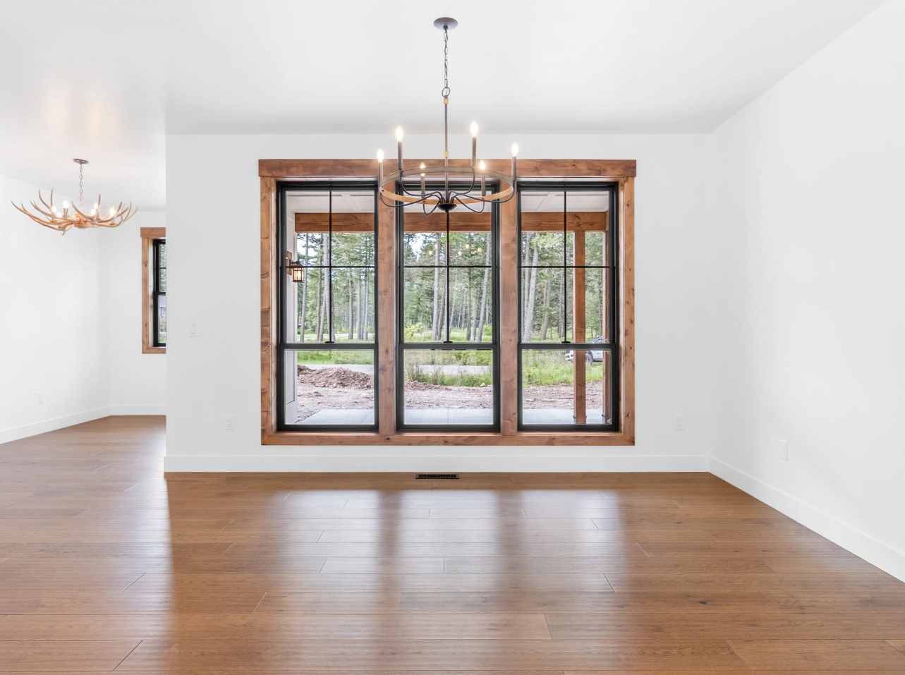 Empty room with wooden floors, large window with black frames and wooden trim, white walls, and a black chandelier with candles hanging from the ceiling.