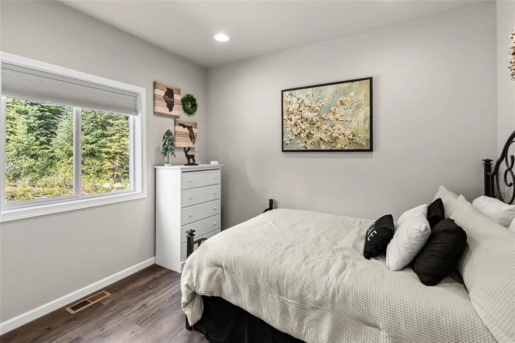 A bedroom with a double bed, black metal headboard, white bedding, and multiple pillows. A window with a white blind showing green trees outside. Decor on the walls includes wooden artwork and a framed painting of cherry blossoms.