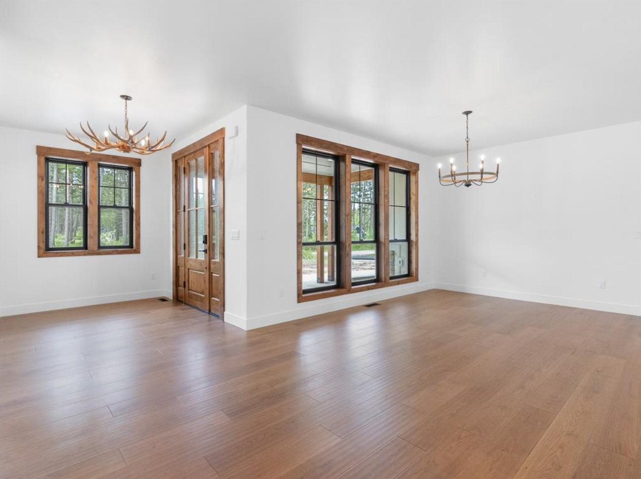 Empty living room with hardwood floors, white walls, large windows with wooden frames, and two chandeliers.