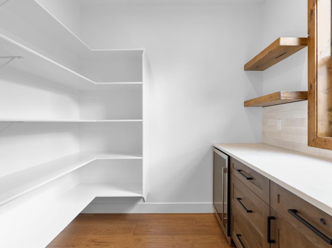 Empty kitchen pantry with white shelves on the left, a countertop with wooden drawers and cabinets on the right, and wooden shelves above on the right side.