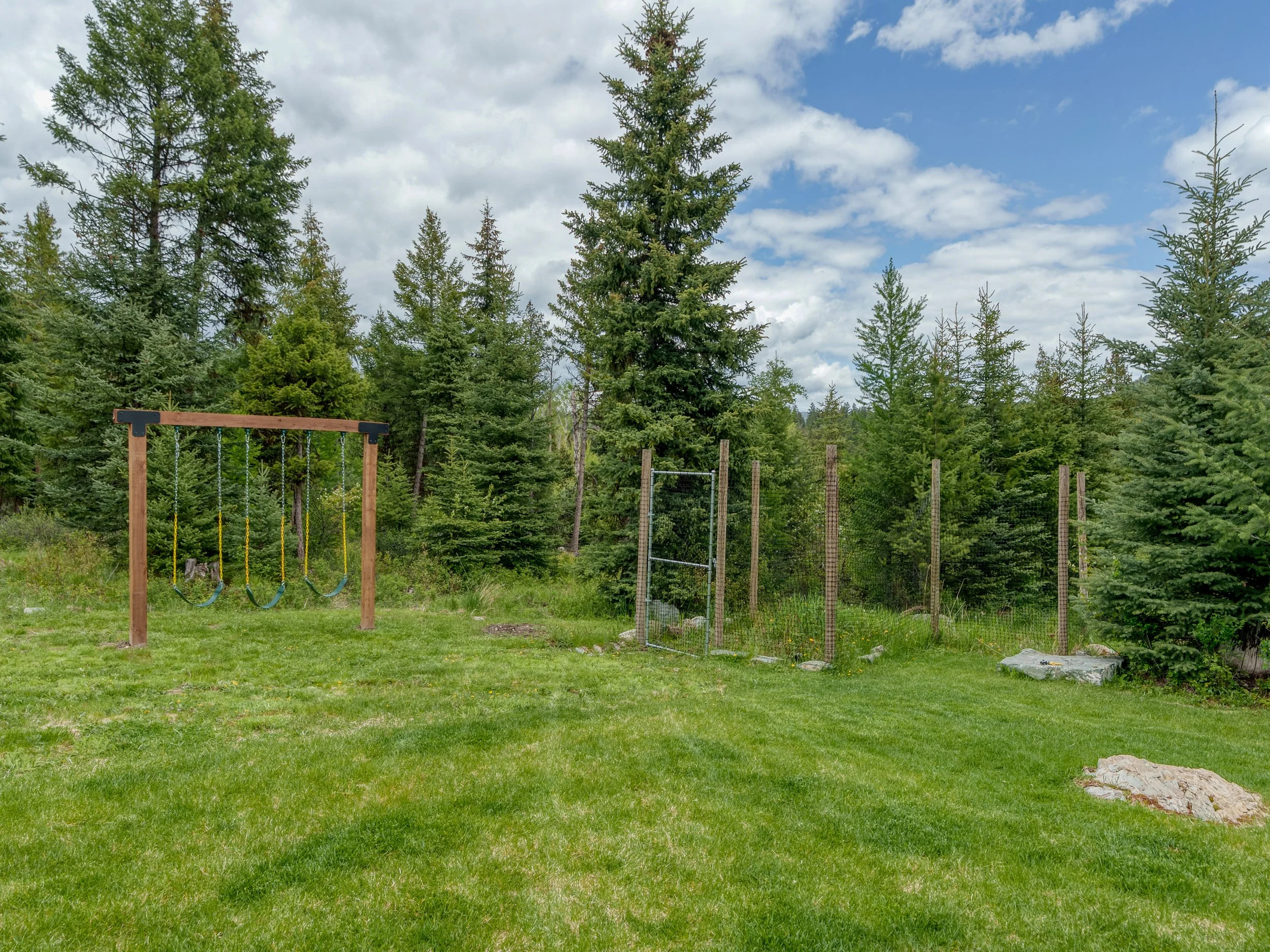 A backyard with lush green grass, surrounded by tall pine trees and a partly cloudy sky, also features a wooden swing set with three swings.