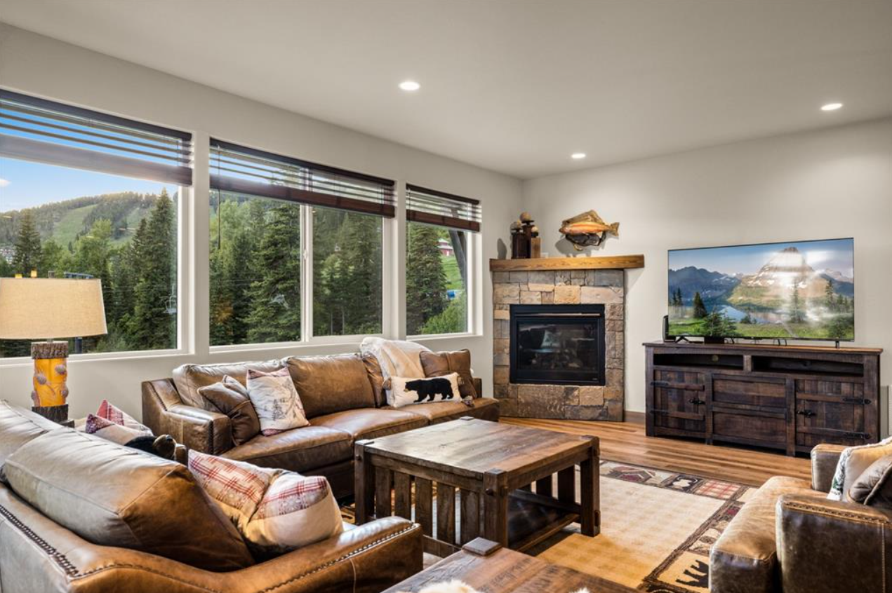Living room with large windows showing a mountain landscape, brown leather sofas with pillows, a fireplace with stone and wood mantel, a wooden coffee table, a flat-screen TV on a wooden stand, lamps, and wooden floors.