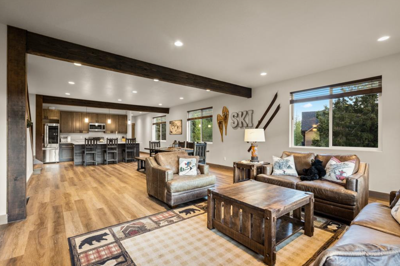 Living room with wood floors, beige leather sofas, wooden coffee table, and table with lamp. Large window showing trees outside. Art on the wall including skis and wooden letters 'SKI'. Kitchen with island, dark stools, and stainless steel appliances