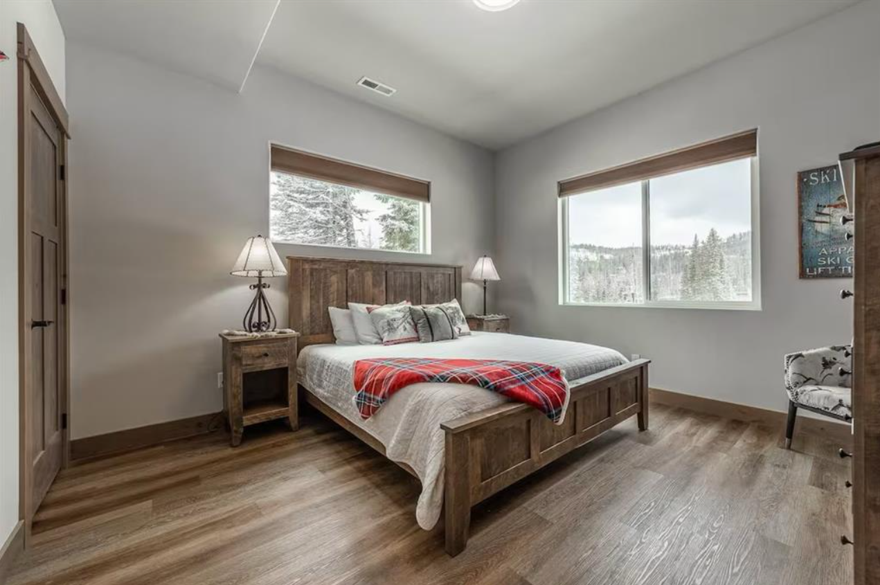 A bedroom with a wooden bed frame, a white comforter, and pillows, flanked by two nightstands with lamps, with a large window showing a snowy landscape, and a wooden wardrobe on the left.