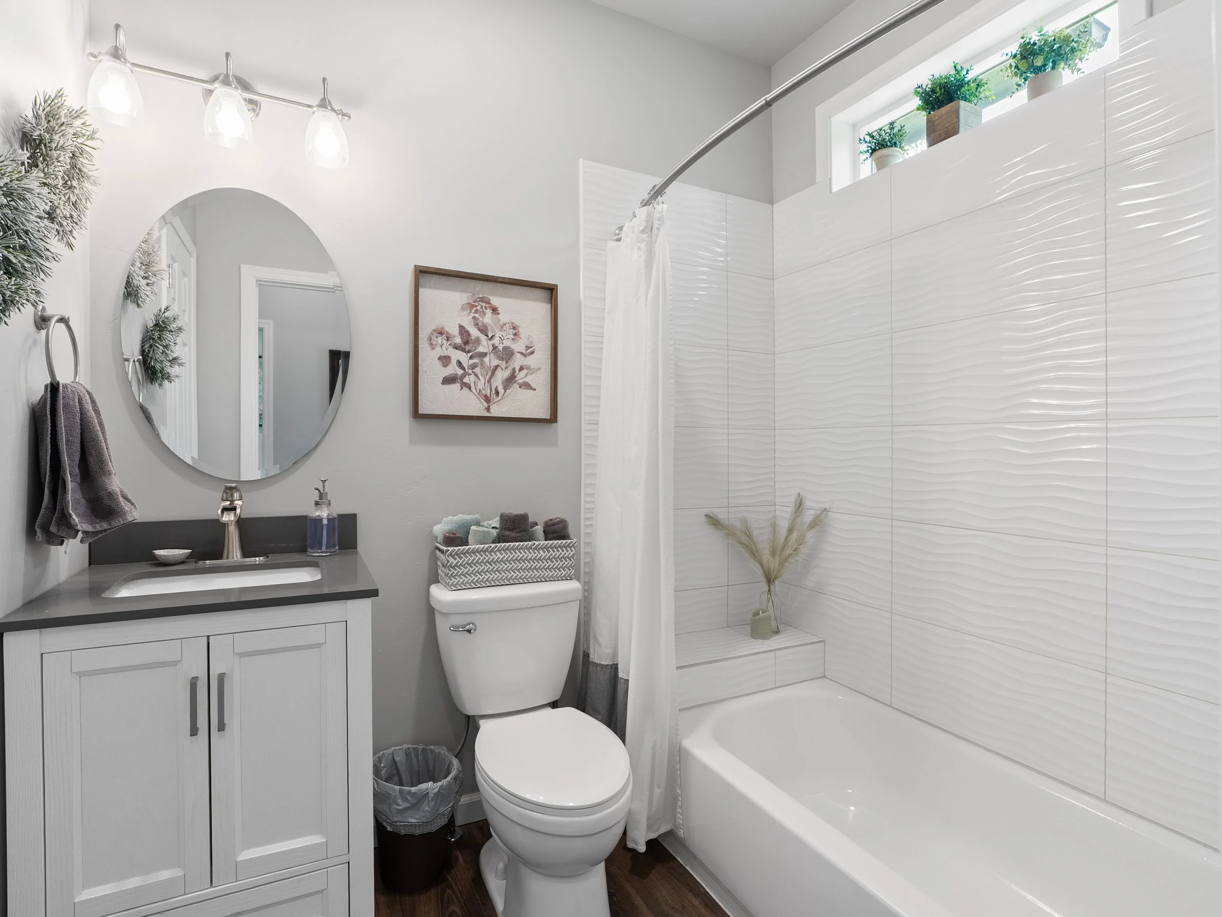 A small bathroom with white walls and a large walk-in shower with textured white tiles and a window with potted plants. There is a white toilet, a vanity with a gray countertop, a mirror, and a cabinet below. Towels are hanging on metal rings, and de