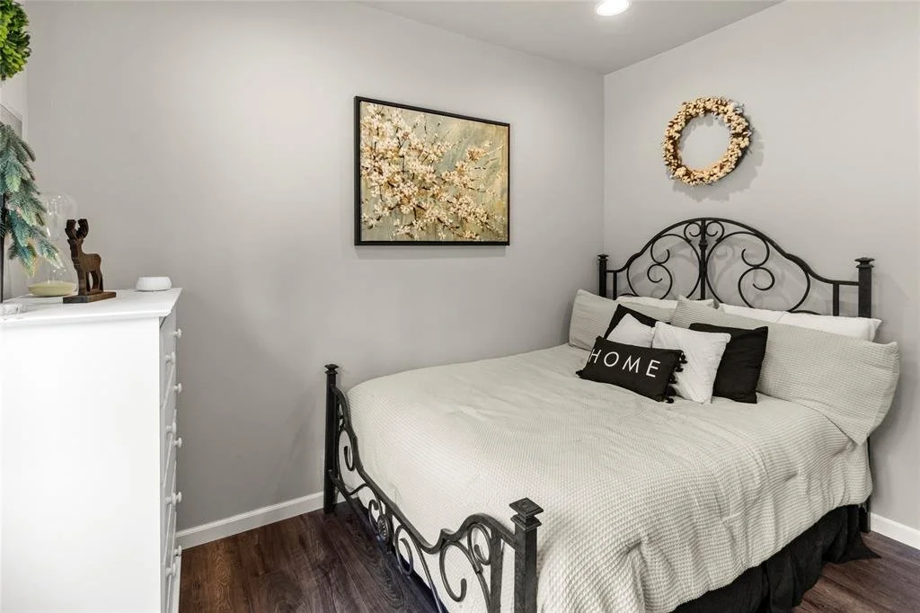 A cozy bedroom with a black metal bed frame, white bedding, and decorative pillows, including one with the word 'HOME'. The wall has a framed floral artwork and a wreath. There is a white dresser with small decorative items, and dark wood flooring.
