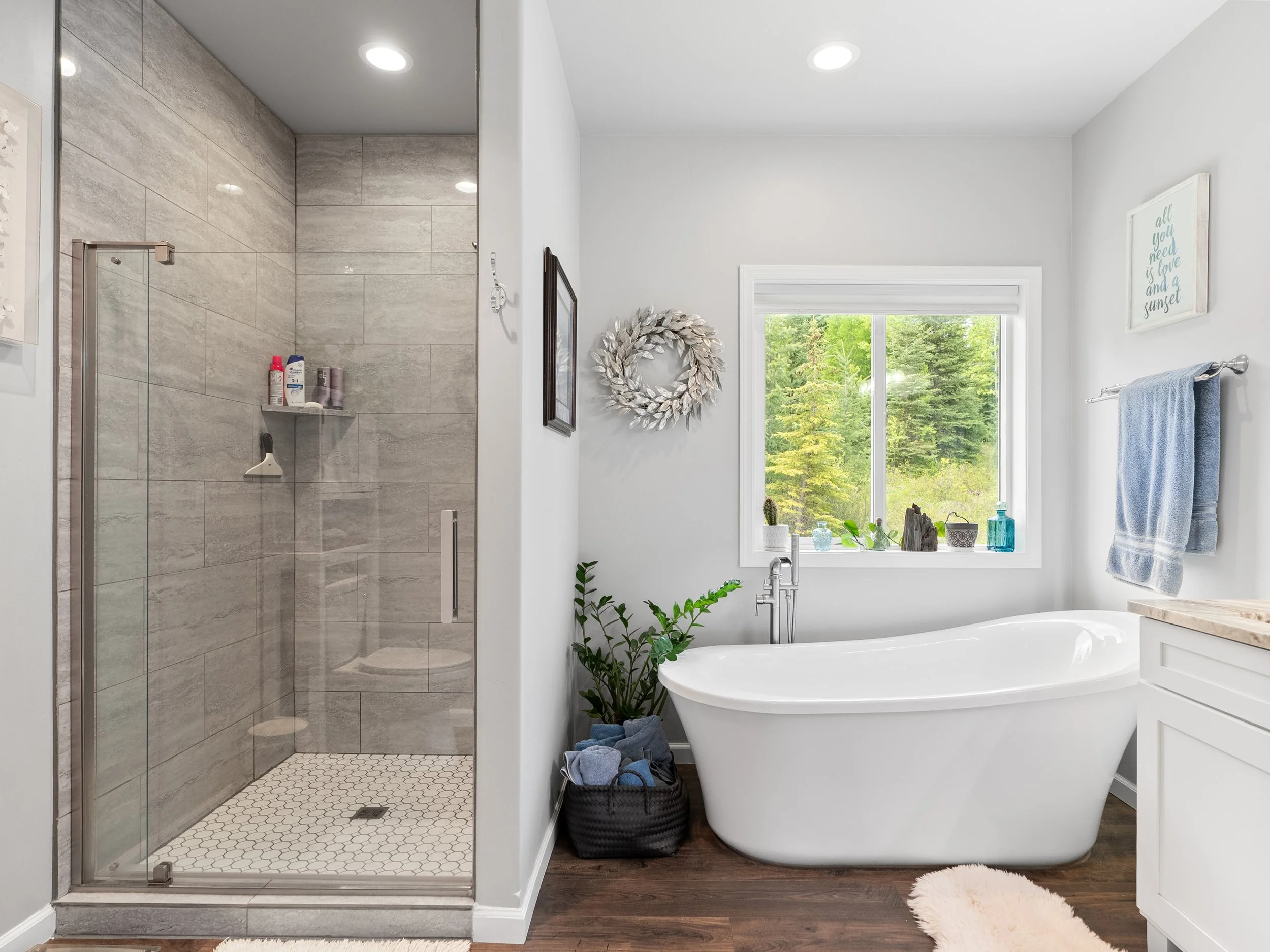 A modern bathroom with a glass-enclosed shower on the left and a white freestanding bathtub near a window on the right. Decor includes a round wreath on the wall, framed art, plants, and towels.