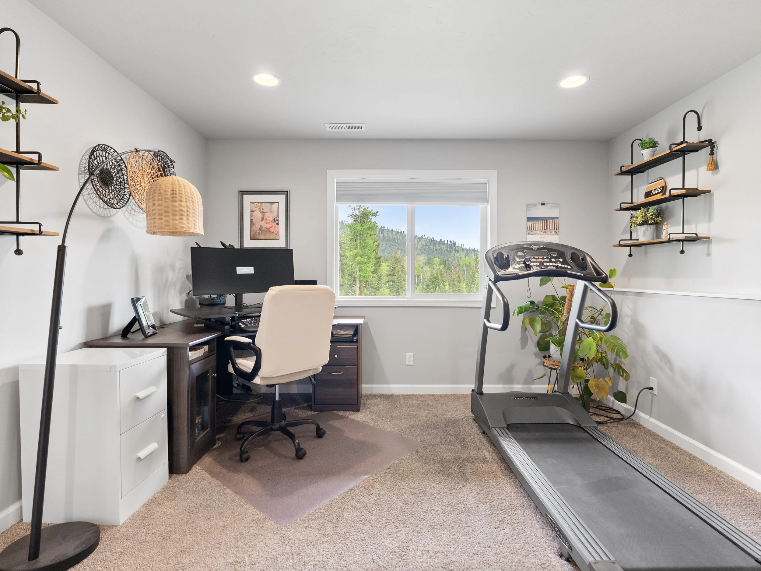 A home office with a treadmill, a desk with a computer, and wall shelves with plants and decorations, overlooking a window with scenic mountains and trees outside.