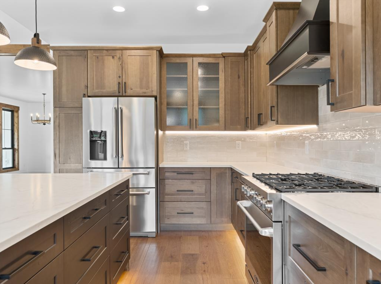 Modern kitchen with wooden cabinets, a stainless steel refrigerator, a gas stove, and a white countertop. Recessed lighting and pendant lights are also visible.