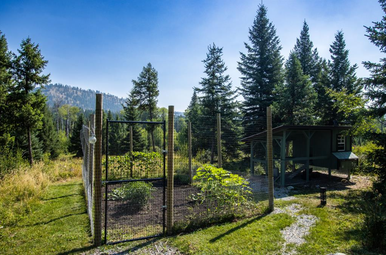 A small garden enclosed with wire fencing in a forested area with trees and hills in the background, and a small playhouse nearby.