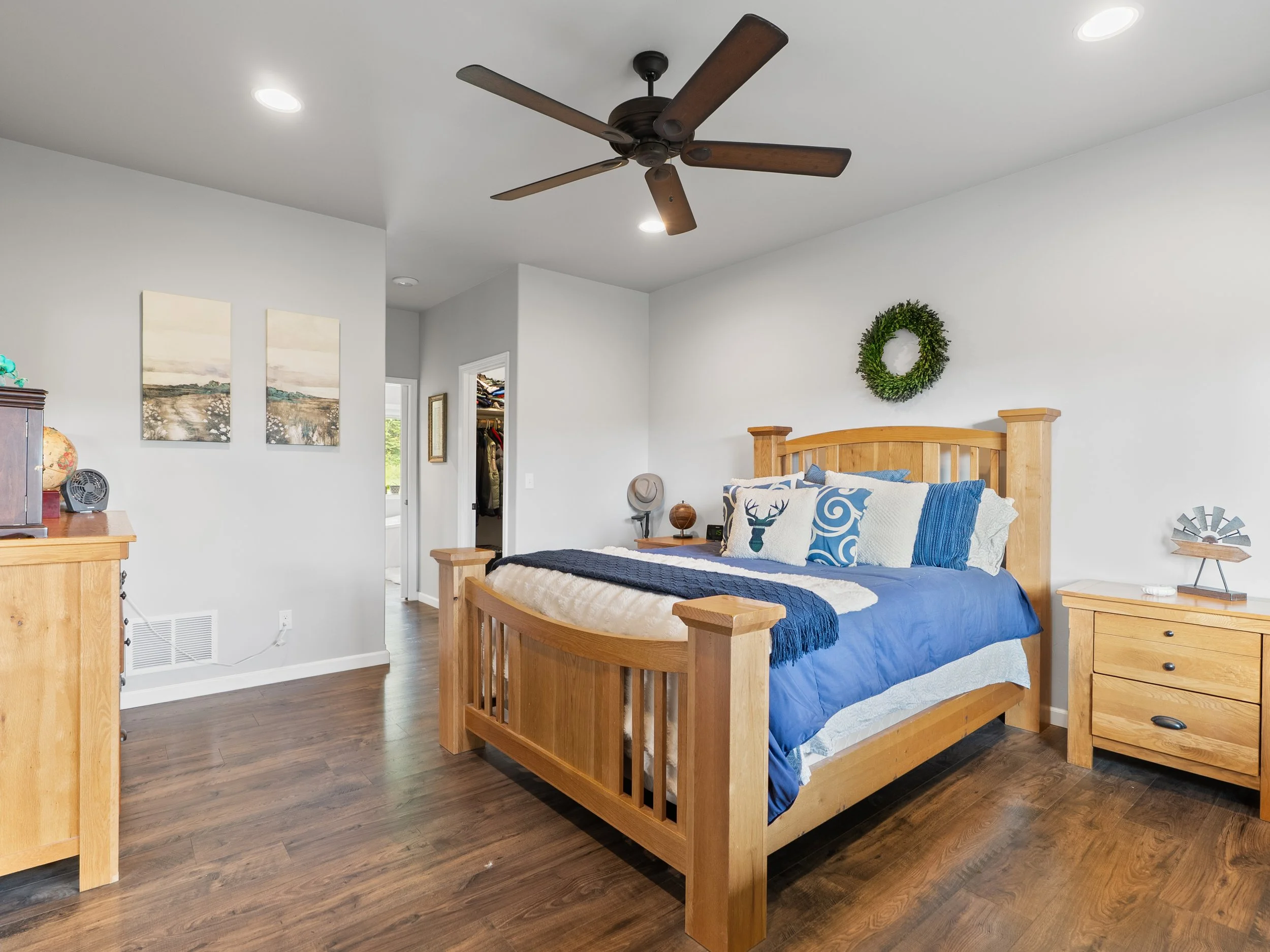 A bedroom with a wooden bed frame, blue bedding, and decorative pillows featuring a deer and swirl design. A green wreath hangs on the white wall above the bed. There are two wooden nightstands, with a hat on the right one. On the left, part of a woo
