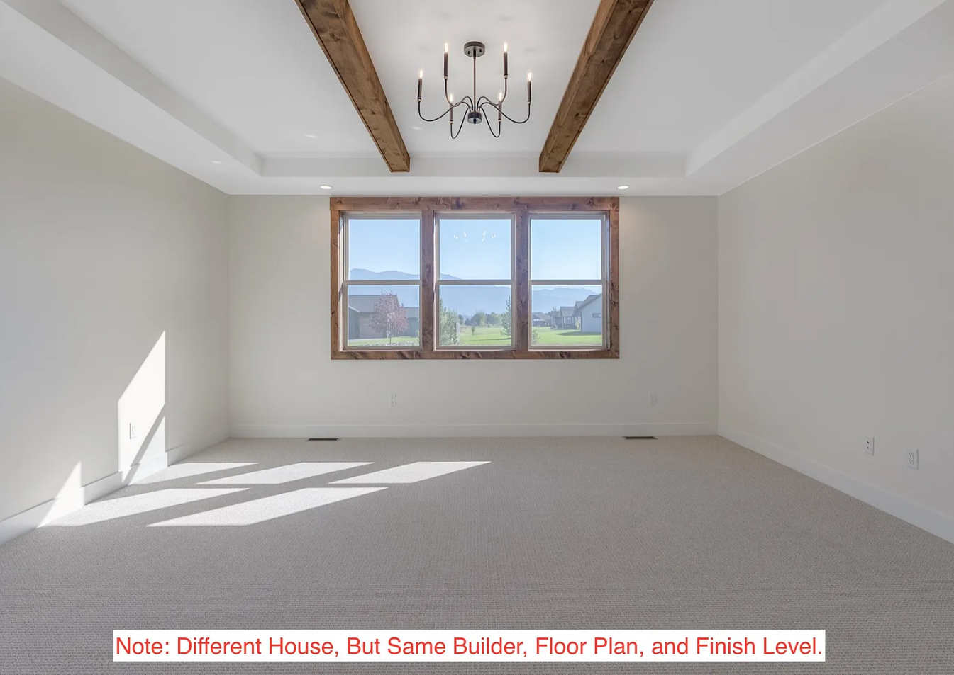 Empty living room with white walls, beige carpet, large window with wooden trim, and wooden beams on the ceiling, lit by natural light and a chandelier.