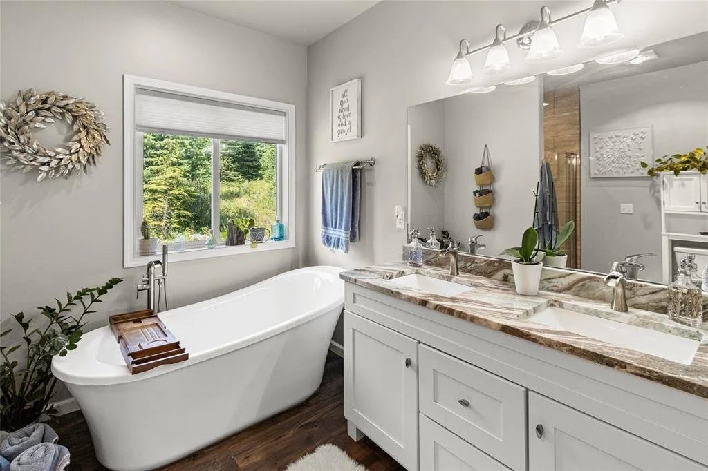 Bathroom with a white bathtub, a large mirror over a marble countertop with two sinks, a window with a view of greenery, and various decorations including a wreath, plants, and framed art.