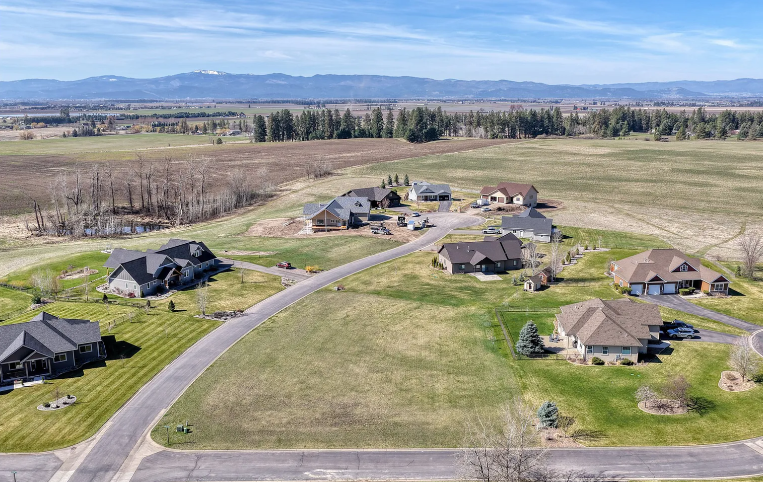 Aerial view of a suburban neighborhood with multiple houses, green lawns, and a curved road, with open fields and distant mountains in the background.
