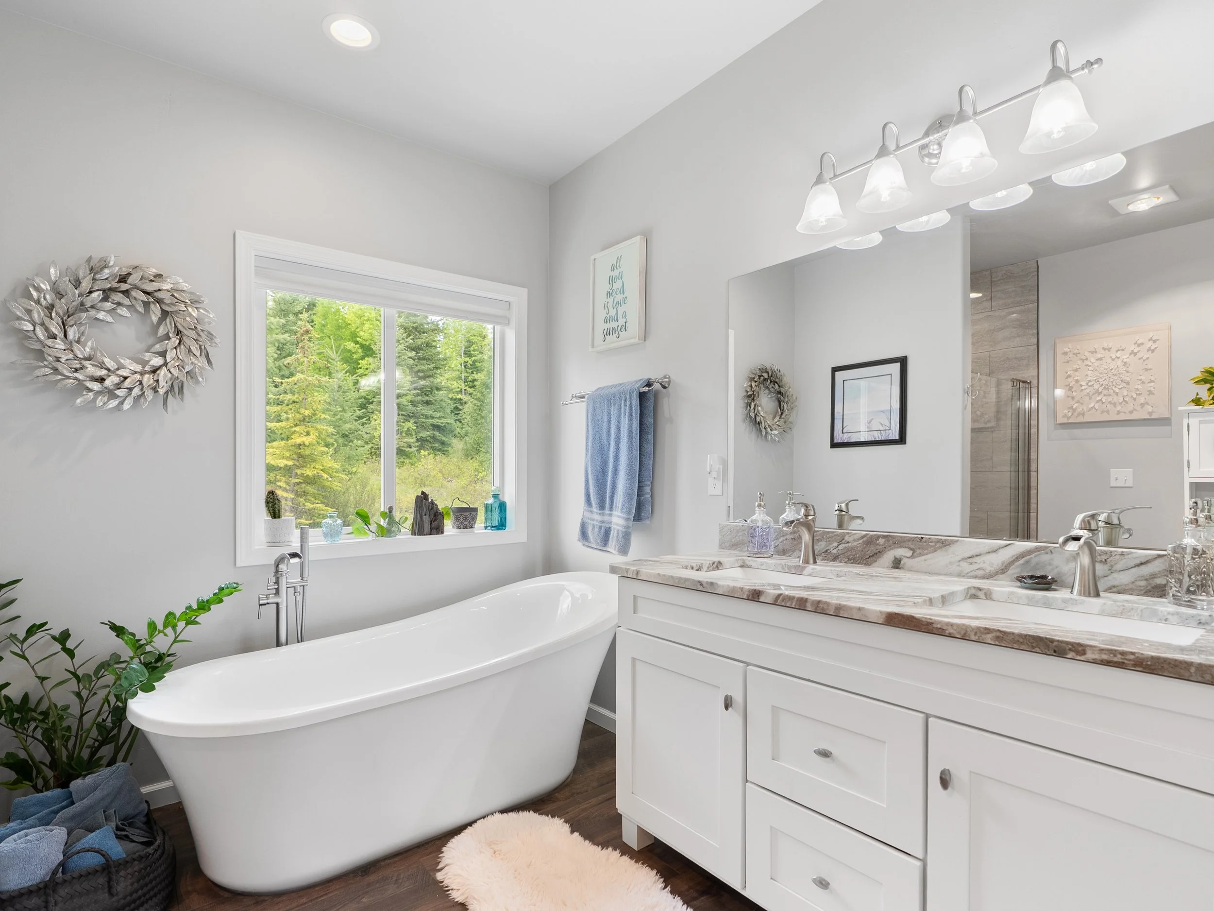A bright bathroom featuring a freestanding bathtub beneath a window with greenery outside. Decor includes a metallic wreath on the wall, framed art, and plants on the window sill. A vanity with a marble countertop and double sinks, framed by a large 
