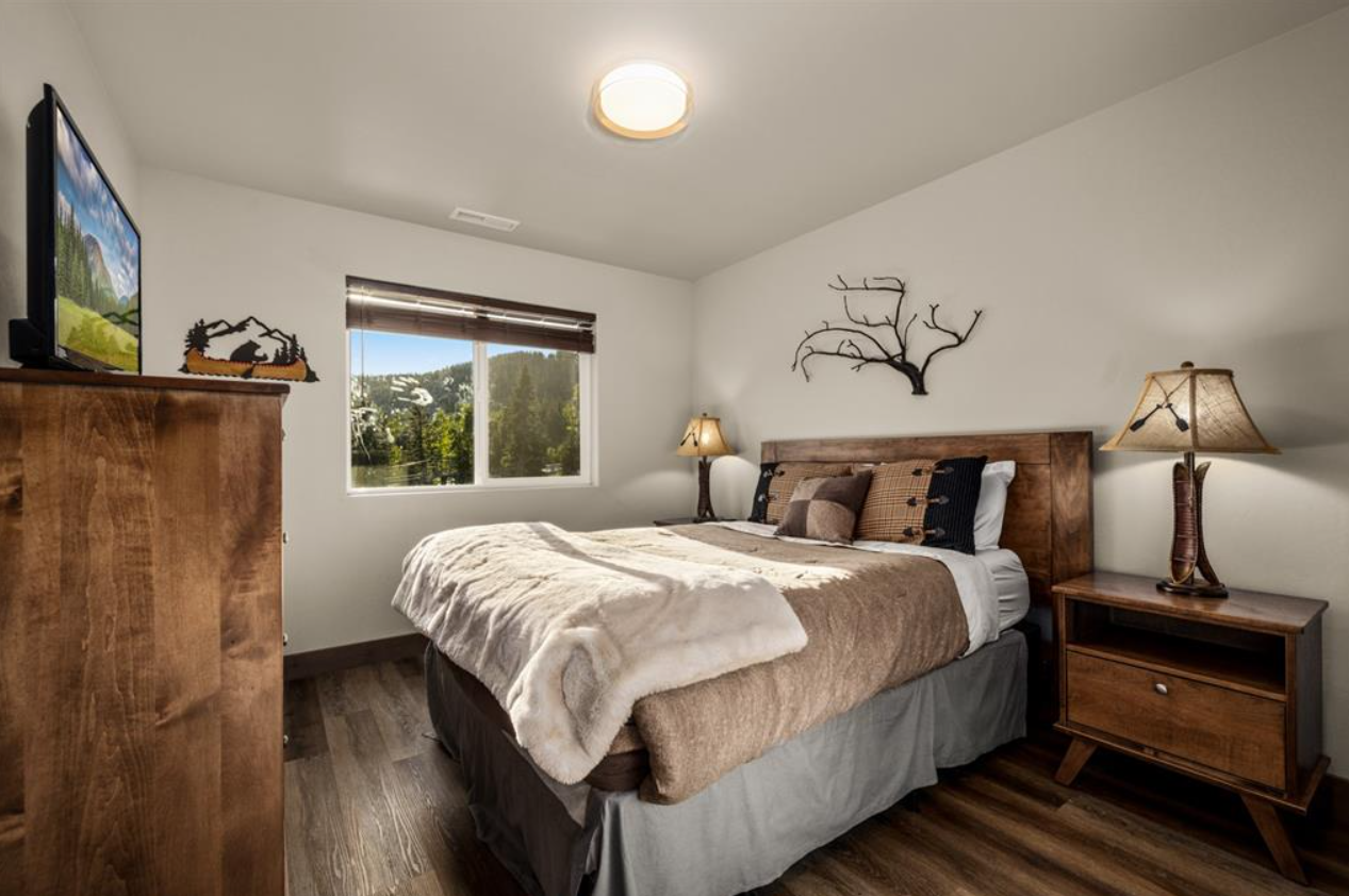 A cozy bedroom with a wooden bed frame, beige bedding, and decorative pillows. There are two wooden nightstands with lamps on each side, a dresser with a TV and decorative items, a window with a view of trees and hills, and a wall art piece resemblin