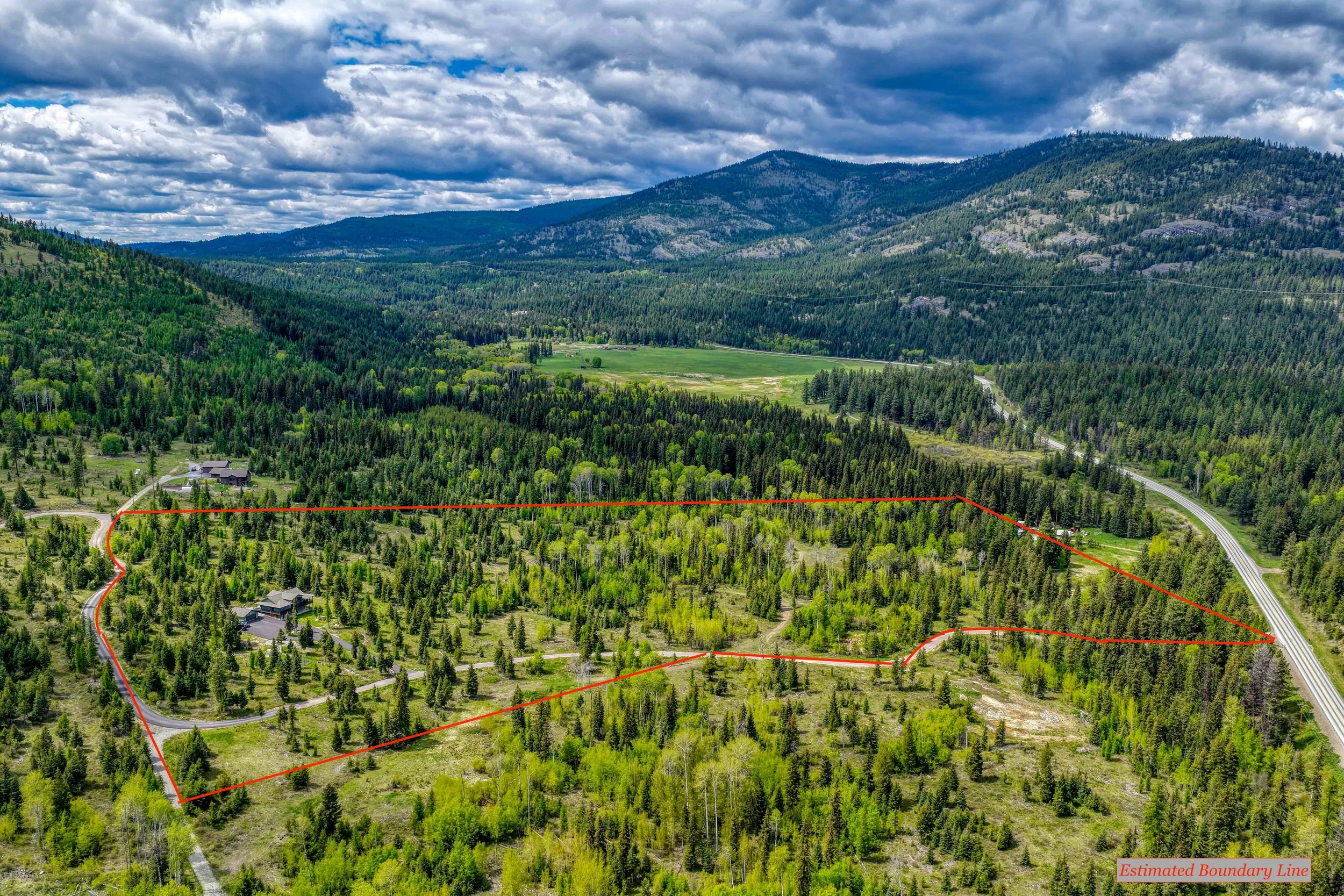 Aerial view of a mountainous landscape with a mix of dense green forests and open grassy areas, under a partly cloudy sky, featuring a marked boundary line in red and a small house at the bottom left corner.