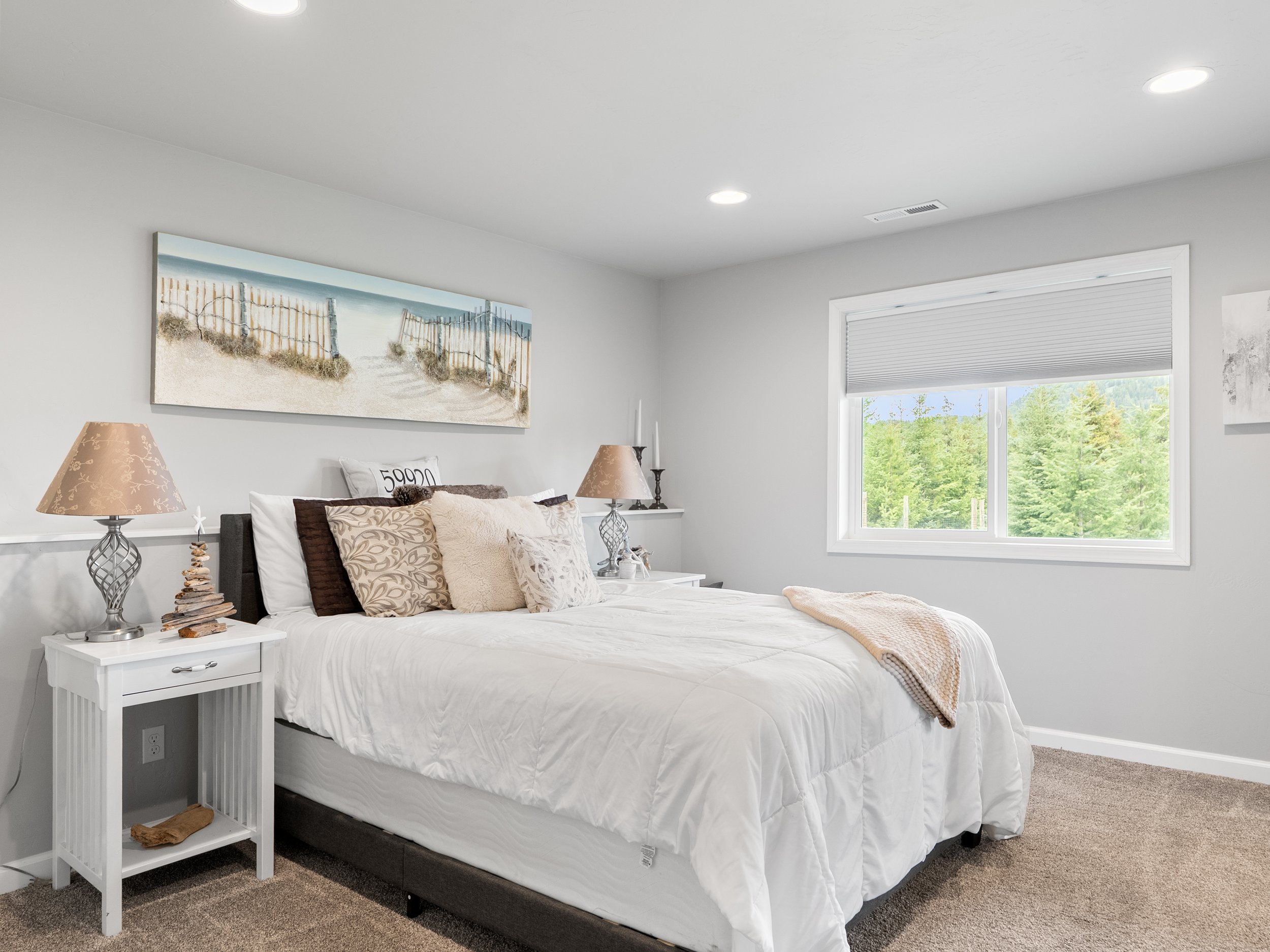 A bedroom with a window showing greenery outside. The bed has white bedding with various pillows, and a blanket at the foot. On the left side, there is a white nightstand with a coral-shaped table lamp and decorative items. Above the bed hangs a land