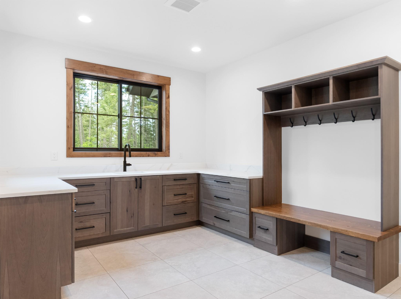 Empty mudroom with gray wooden cabinets, white marble countertops, a black faucet, a large window with wooden trim, and wall hooks for coats.