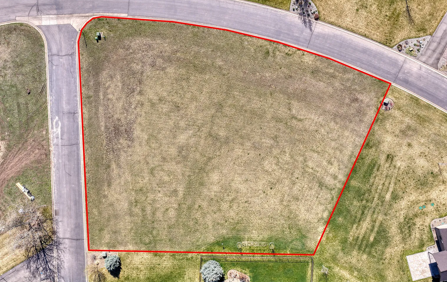 Aerial view of a vacant, grassy lot outlined in red, surrounded by roads and some trees, with neighboring houses visible.