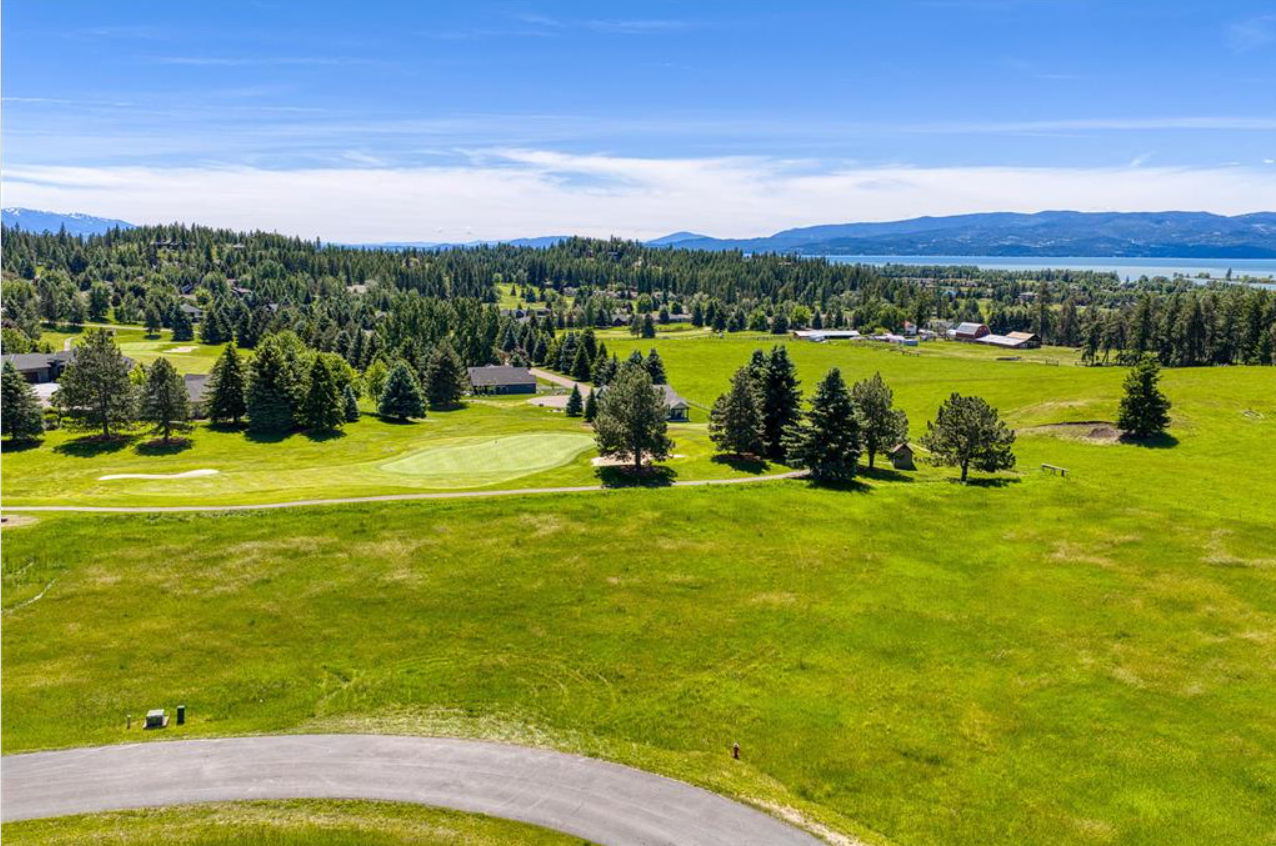 A scenic landscape with a golf course, lush green grass, trees, houses, and a distant mountain range under a blue sky.