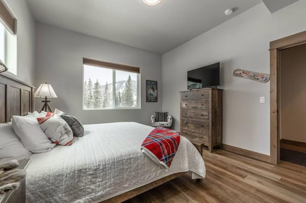 A cozy bedroom with a large bed covered in white bedding, a wooden headboard, and multiple pillows. There is a window showing snow-covered trees outside, a side table with a lamp, a patterned chair, a wooden dresser with a television on top, and a de