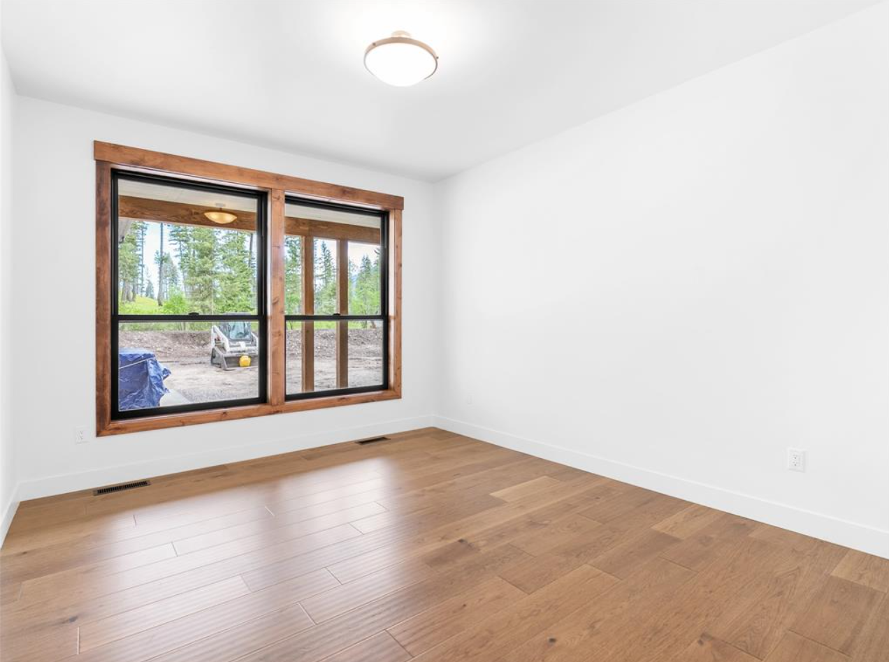 Empty room with white walls, wooden floor, and a large window with wood trim overlooking outdoor trees and patio area.