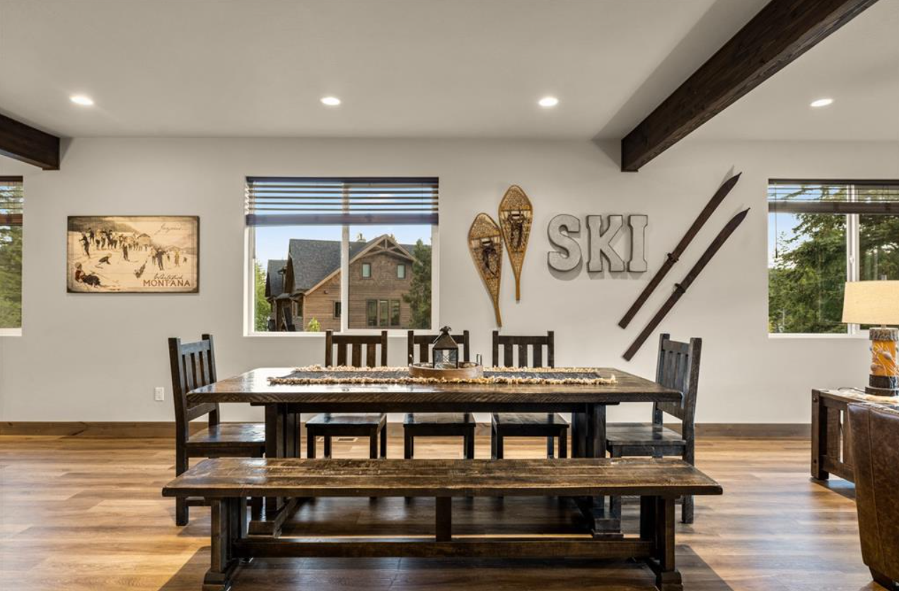 Interior of a dining room with a dark wooden table, matching chairs, window views of houses and trees, and wall decorations including ski-themed ornaments and a Montana poster.