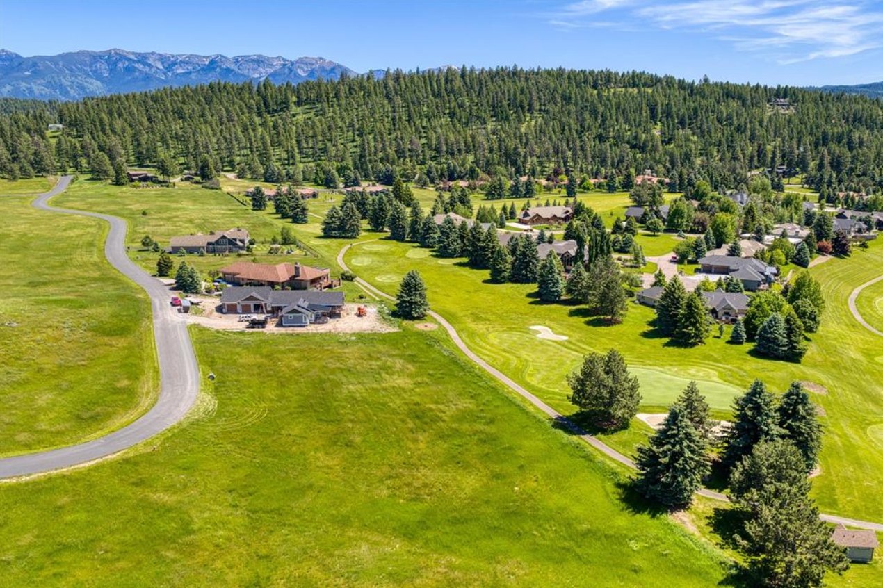 Aerial view of a lush green golf course with several houses and trees, against a backdrop of mountains and a blue sky.