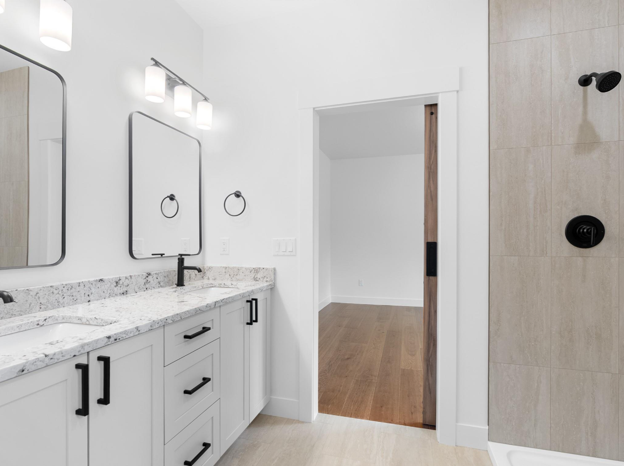 Modern bathroom with double vanity sinks, granite countertops, black fixtures, large mirrors, white walls, towel rings, wall lighting, and a walk-in shower with beige tiles and black fixtures.