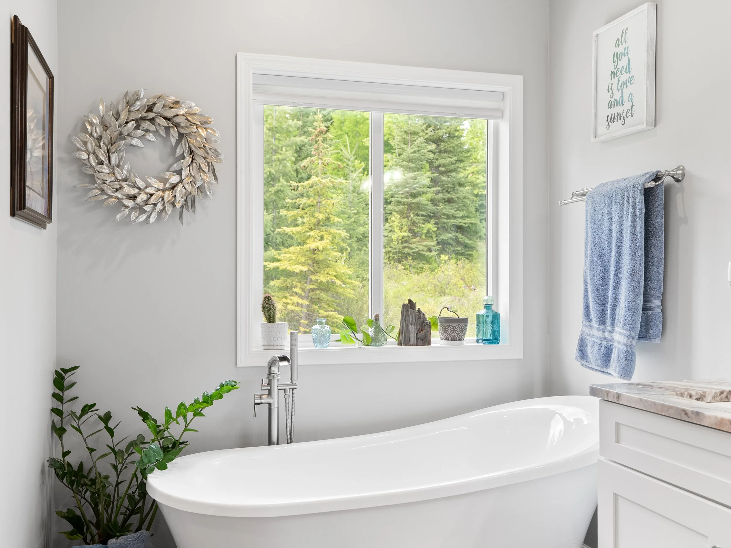 A bathroom with a white bathtub, green plants, decorative bottles, a window with greenery outside, and wall decor including a wreath and a framed quote.