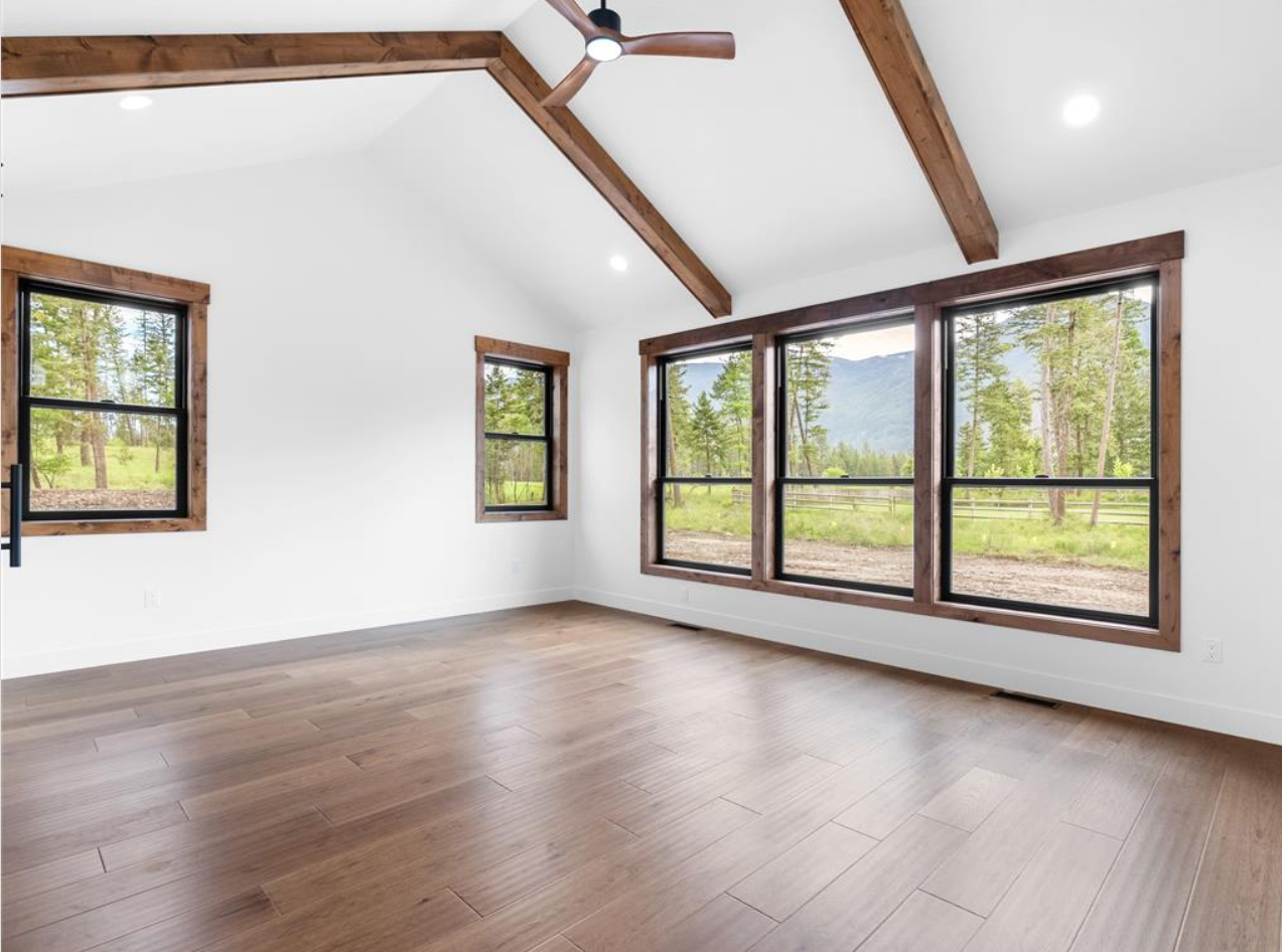 An empty room with large windows showing a scenic outdoor view of trees and mountains. The room has white walls, wooden trim around the windows, light-colored hardwood floors, and exposed wooden beams on the ceiling. A modern ceiling fan is mounted o