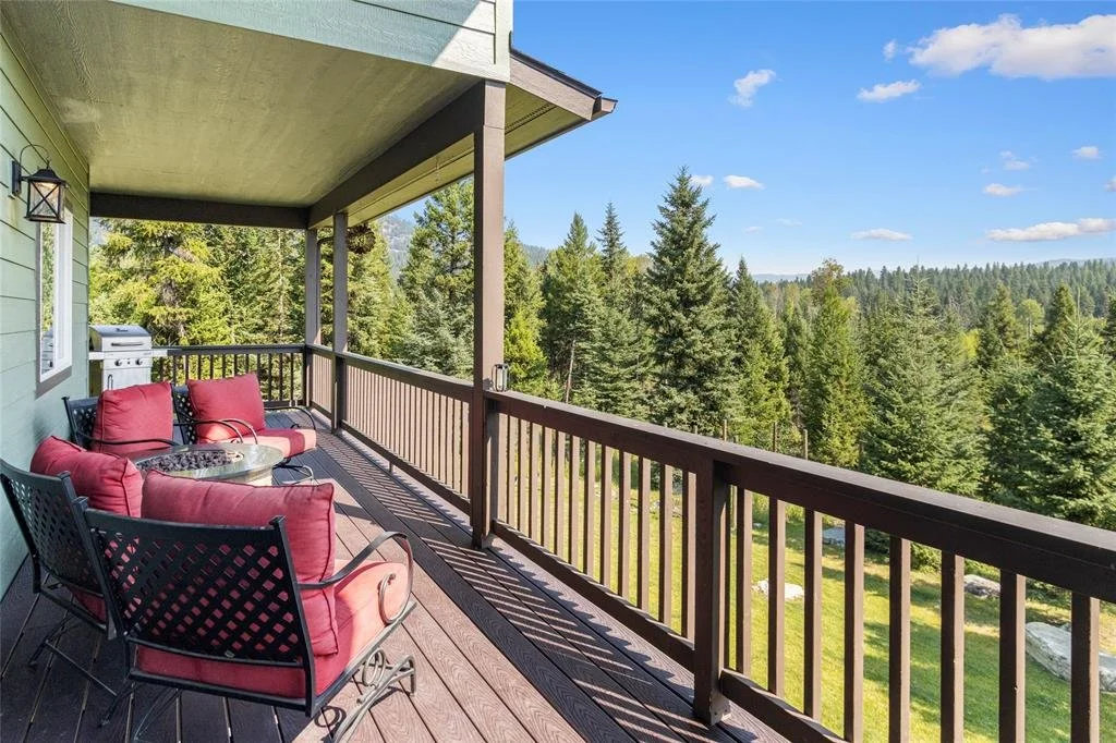 A spacious balcony with outdoor seating, including a sofa and chairs with red cushions, overlooking a lush green forest with tall pine trees under a cloudy blue sky.