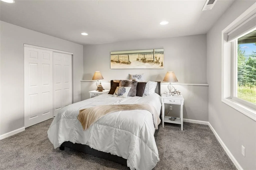 A neatly made bed with white bedding and decorative pillows in a bright bedroom with a window showing green trees outside. Two nightstands with lamps flank the bed, and a beach-themed artwork hangs above it.