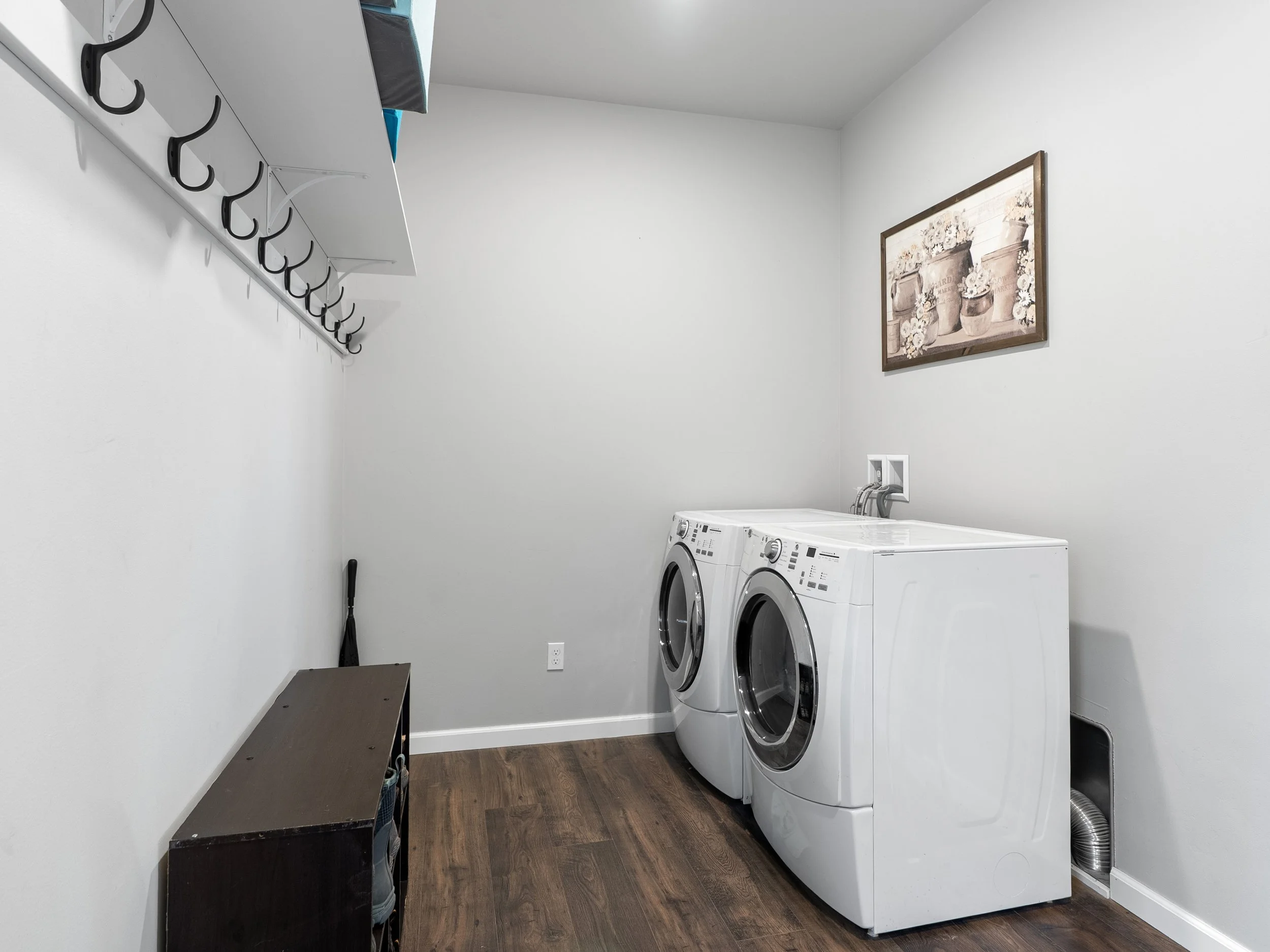 A laundry room with two front-loading washing machines side by side, a dark wooden bench on the left, a picture of potted flowers on the wall, a white wall, and a wooden floor.