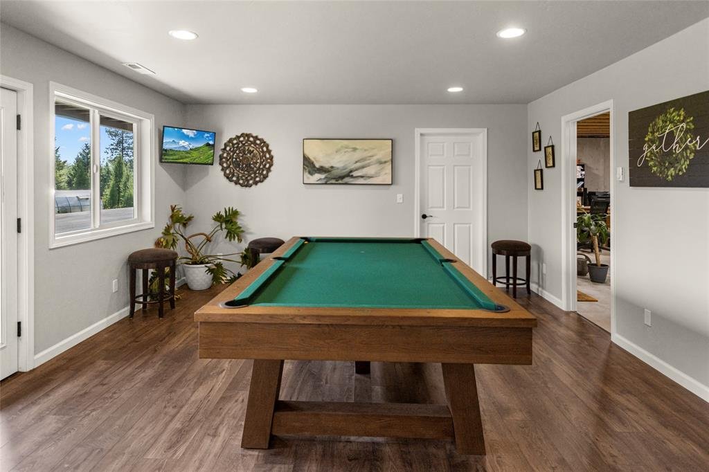 A game room with a pool table in the center, hardwood floors, a window showing a view of trees, wall art, and a small flat-screen TV.