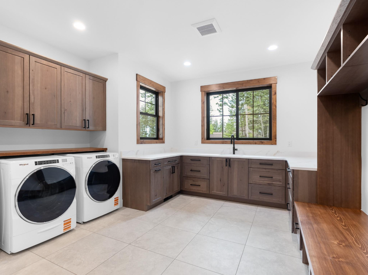 Laundry room with white washer and dryer, brown wooden cabinets, two large windows, and beige tiled floor.