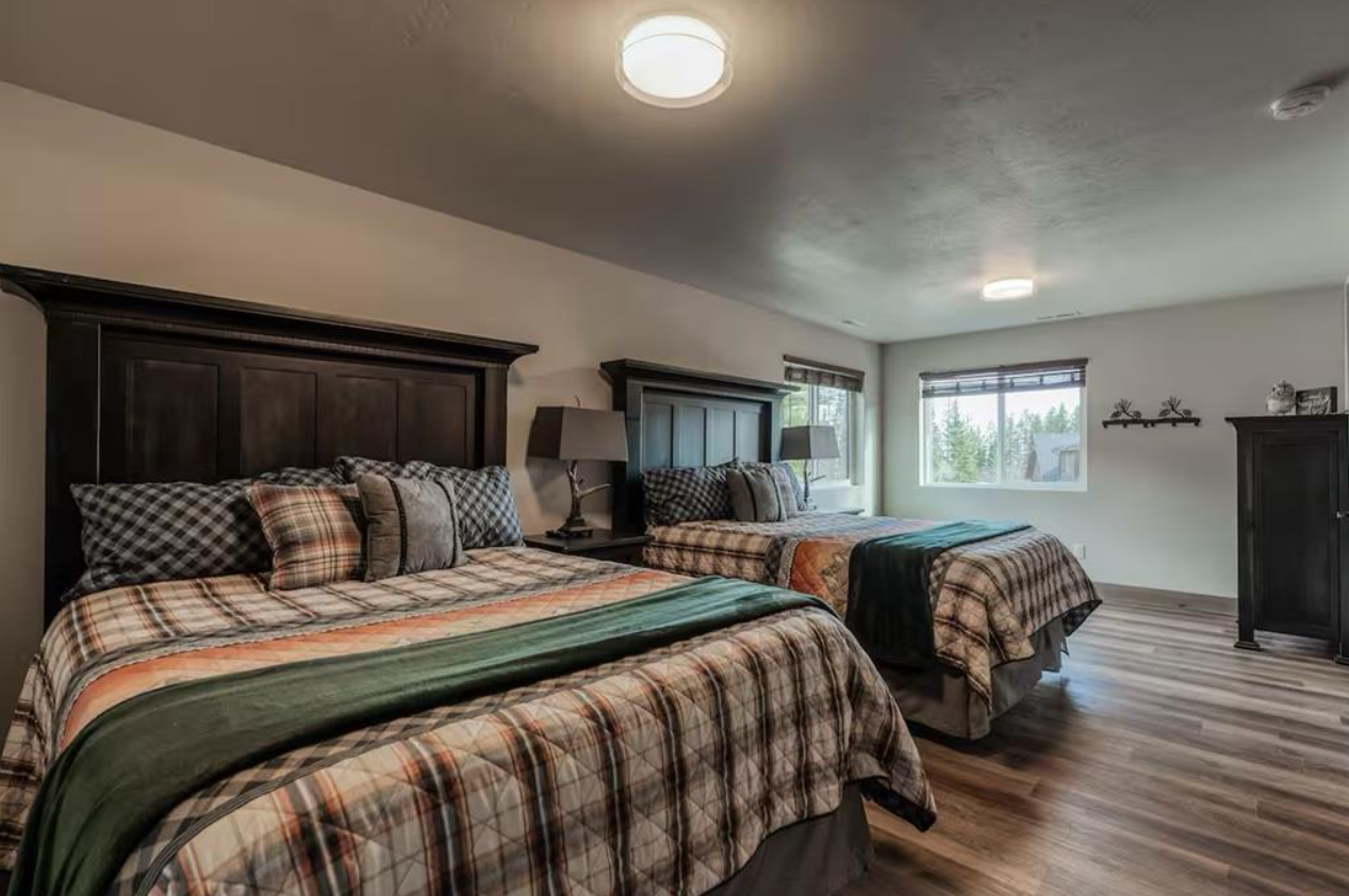 A bedroom with two large beds with plaid bedding and multiple pillows, dark wood headboards, two bedside lamps, wooden floors, and windows with blinds letting in natural light.