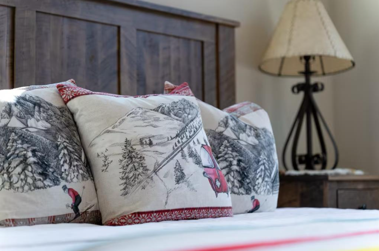 A bed with pillows featuring winter scene patterns, including trees, mountains, and a skier, in a cozy bedroom with wooden furniture and a lamp.