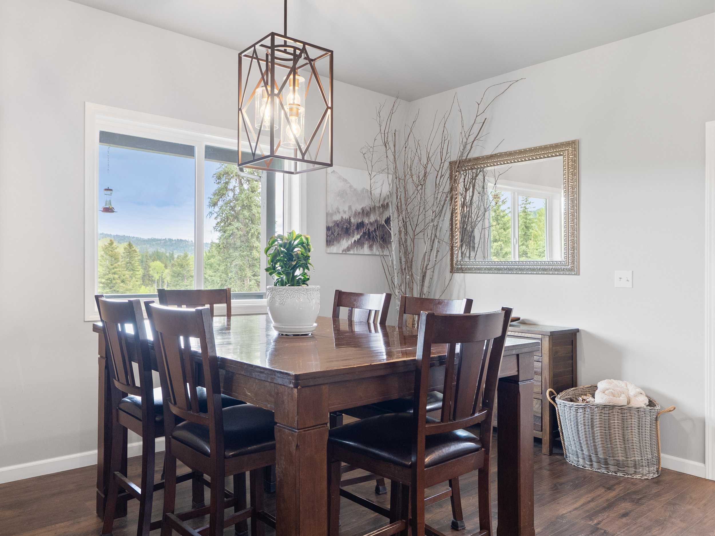 A dining room with a wooden table and six matching chairs, a large window letting in natural light, a potted plant on the table, a framed landscape painting on the wall, a decorative mirror, and a laundry basket in the corner near a side table with t