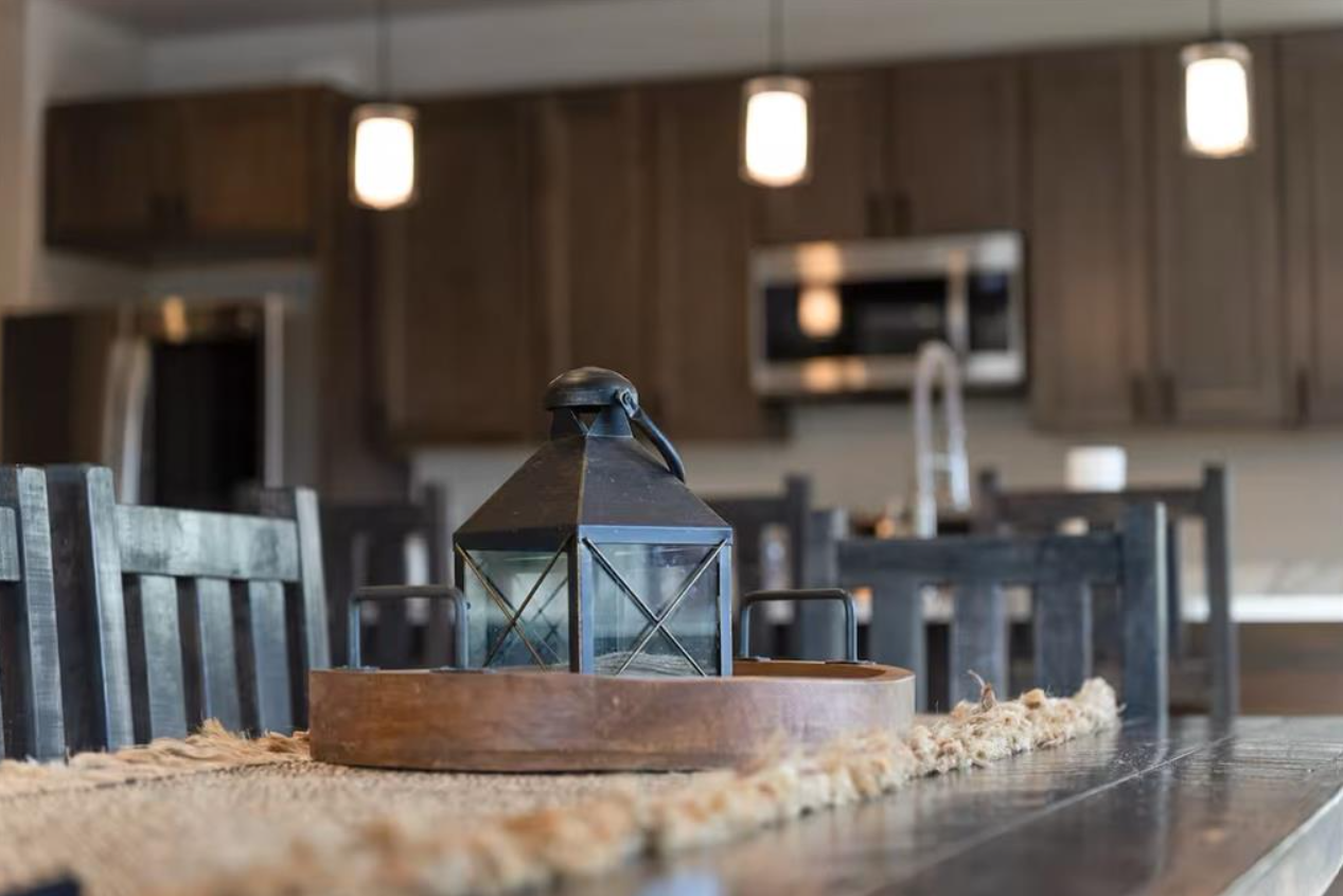 A black lantern on a wooden tray centered on a dining table decorated with a beige table runner, in a modern kitchen with dark wooden cabinets, stainless steel appliances, and pendant lights.