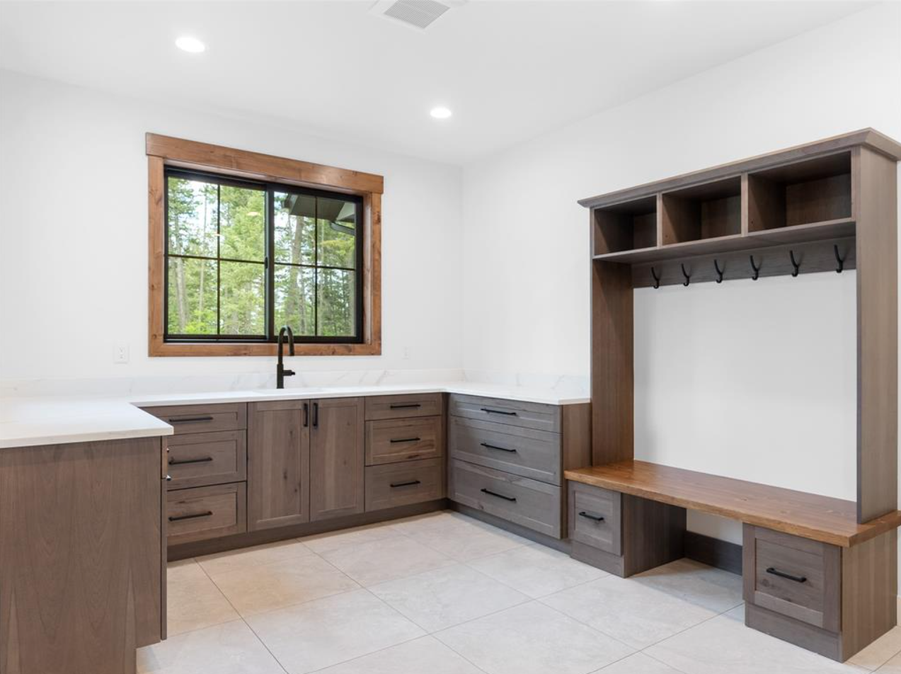 Empty kitchen with wooden cabinets, white countertop, black faucet, window showing trees outside, and built-in wooden bench with coat hooks.