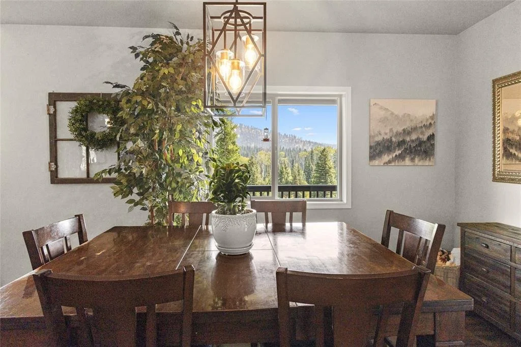 A Dining Room with a wooden table, six matching chairs, a large potted plant, a window showing a scenic mountain view, artwork on the walls, and a modern light fixture overhead.
